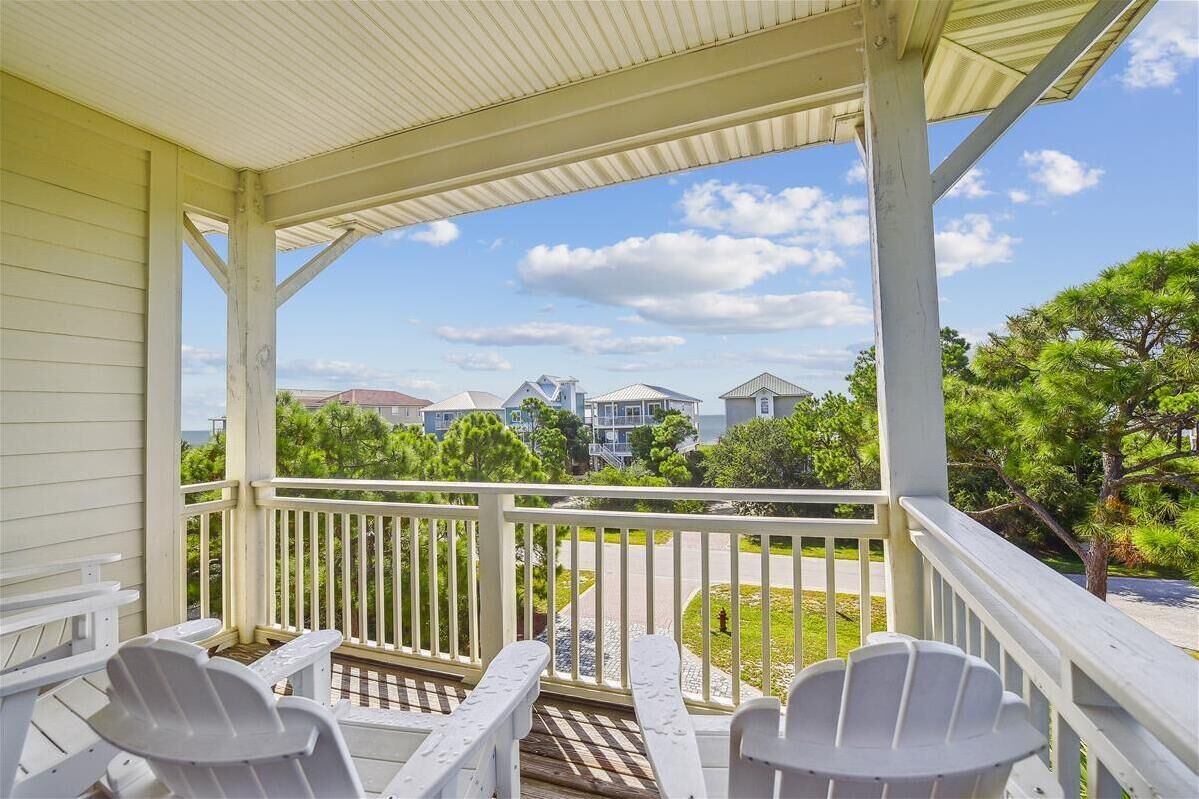 A covered porch with white railings and Adirondack chairs, overlooking a residential street with houses and greenery.