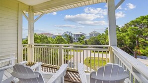 A covered porch with white railings and Adirondack chairs, overlooking a residential street with houses and greenery. - Welcome to Sandy Bottoms! (St. George Island)