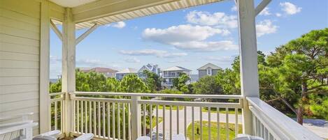A covered porch with white railings and Adirondack chairs, overlooking a residential street with houses and greenery.