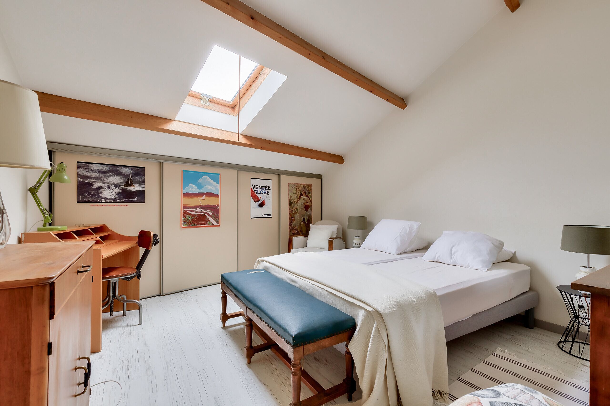 Un dormitorio con cama, banco, escritorio, silla y obras de arte en las paredes.