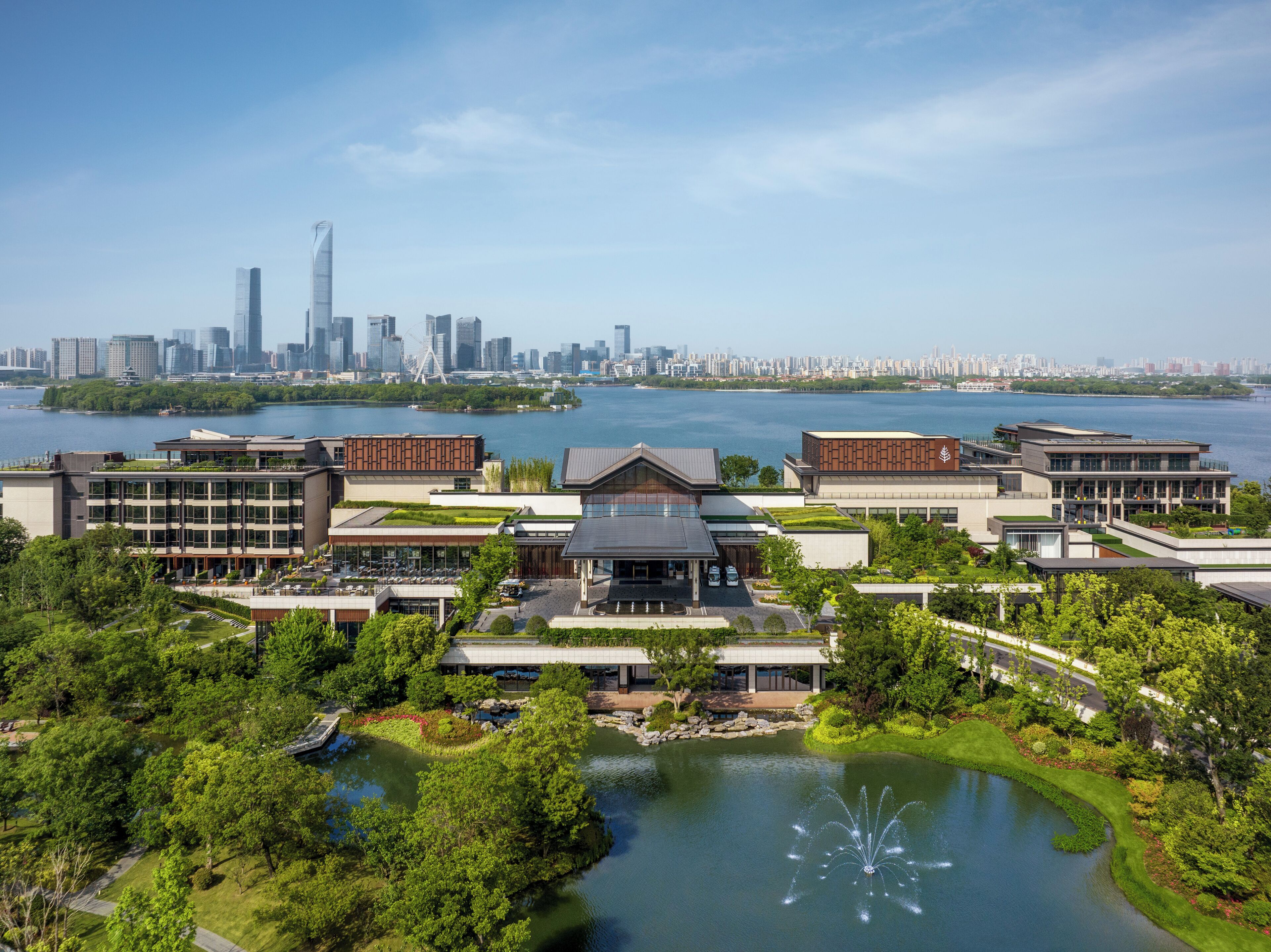 A modern hotel complex with multiple buildings, a central courtyard, and a view of a city skyline across a body of water.