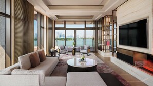 A modern living room with a sofa, coffee table, and chairs, large windows with a view of a cityscape and water, and a flat-screen TV. - Four Seasons Hotel Suzhou (Suzhou)