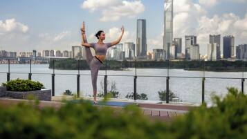 Eine Person führt eine Yoga-Pose auf einem Balkon aus, im Hintergrund ist eine Stadtlandschaft zu sehen.