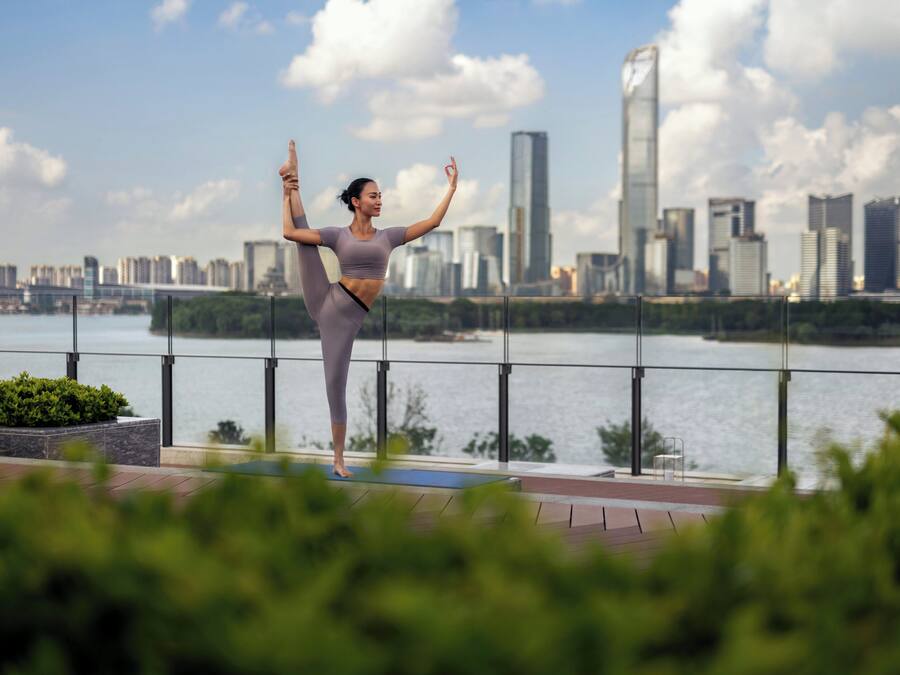 Eine Person führt eine Yoga-Pose auf einem Balkon aus, im Hintergrund ist eine Stadtlandschaft zu sehen.