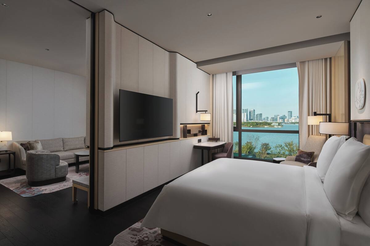 A modern hotel room with a large bed, a flat-screen TV, a seating area with a sofa and armchair, a small table, and a view of the city skyline through large windows.