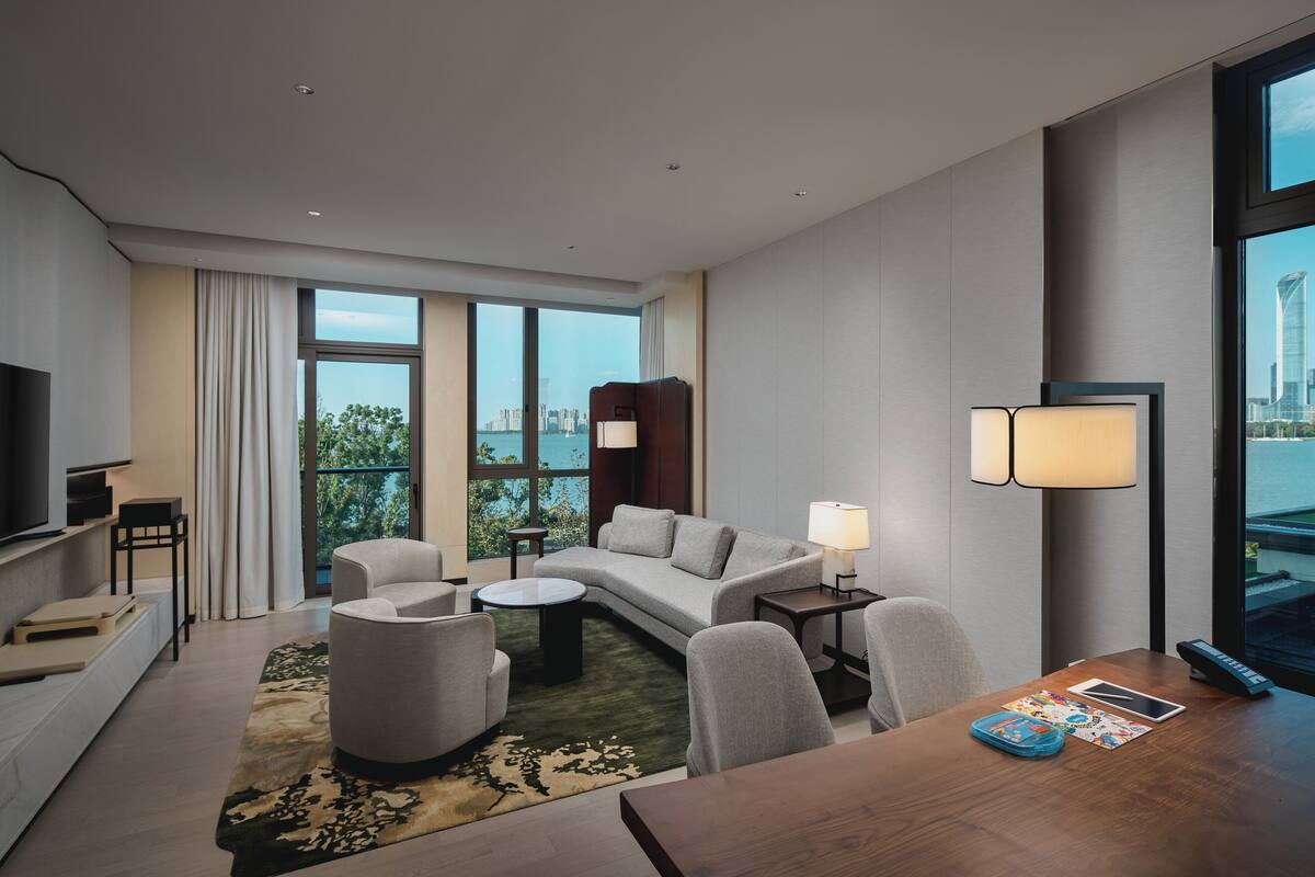 A modern hotel room with a sofa, armchairs, a coffee table, a TV, and a large window offering a city view.