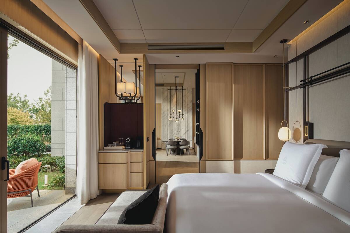 A modern hotel room with a large bed, a desk with a chair, a dining area, and a view of the outdoors.