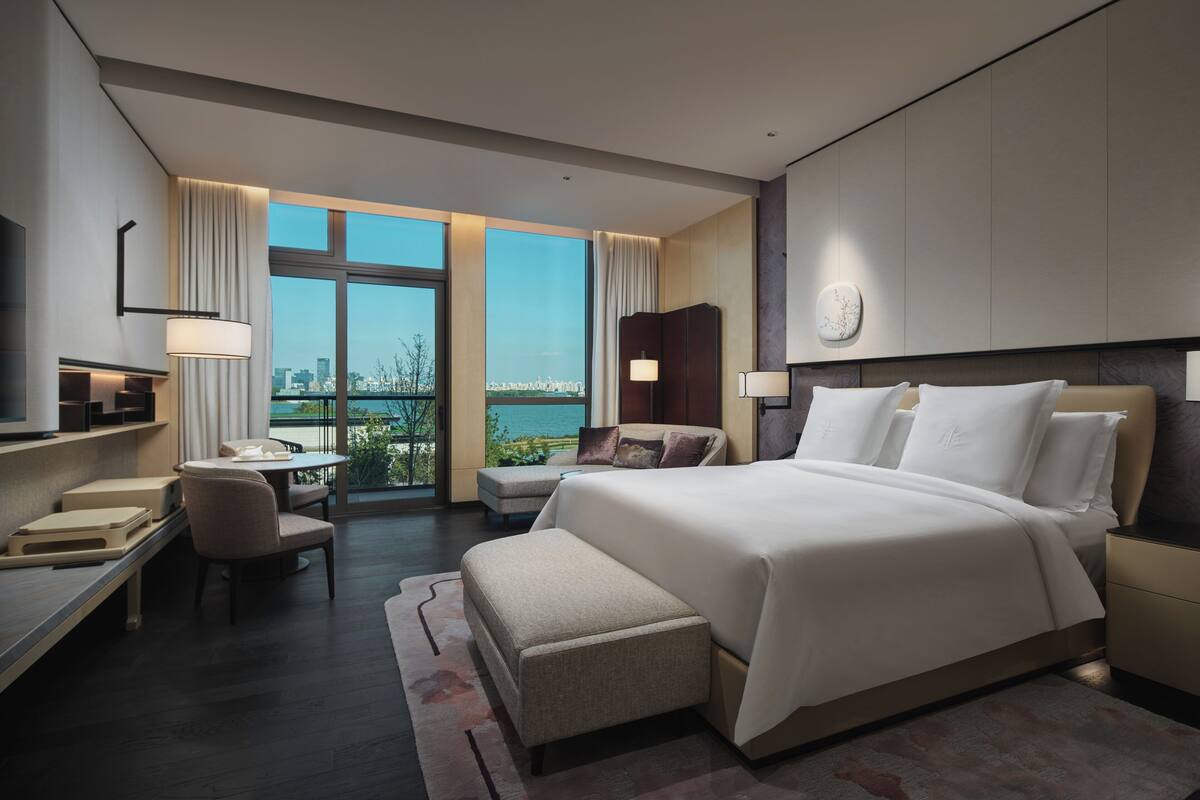 A modern hotel room with a large bed, a desk, a chair, and a view of the city through large windows.