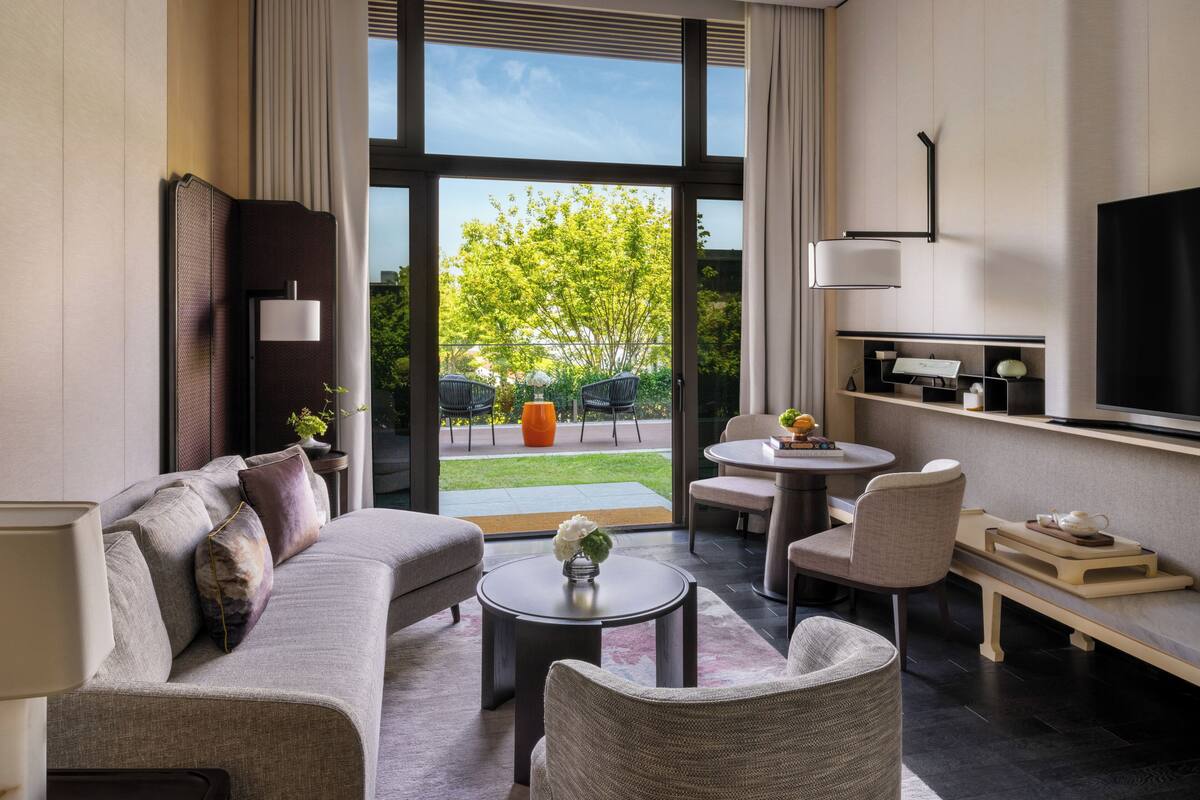 A modern living room with a sofa, coffee table, and chairs, a dining area with a table and chairs, a flat-screen TV, and a view of a garden through large windows.