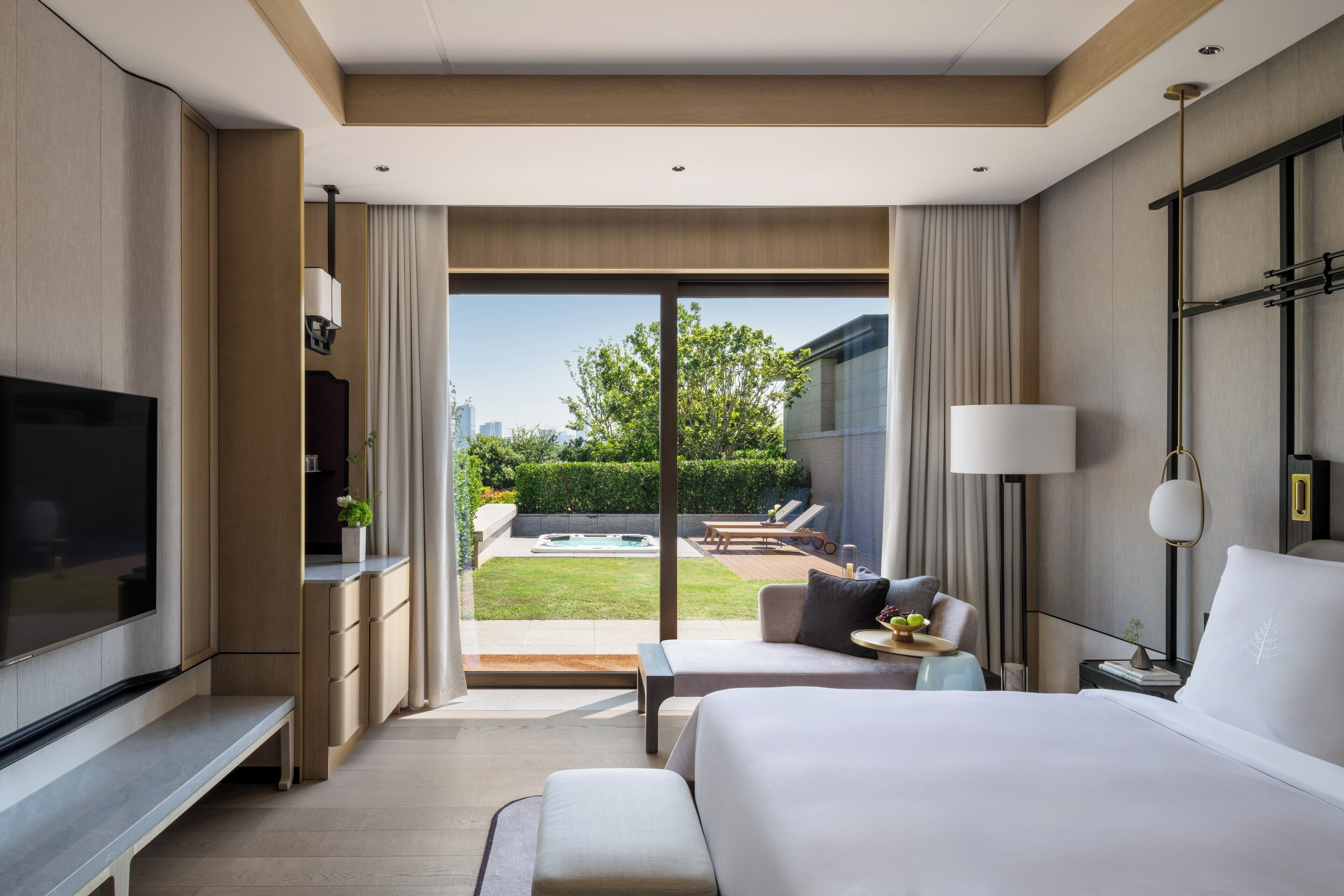 Habitación de hotel moderna con una cama grande, un televisor de pantalla plana, una zona de estar con sofá y sillones, y vistas a un patio con piscina y vegetación.