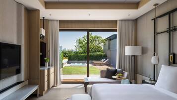 Habitación de hotel moderna con una cama grande, un televisor de pantalla plana, una zona de estar con sofá y sillones, y vistas a un patio con piscina y vegetación.
