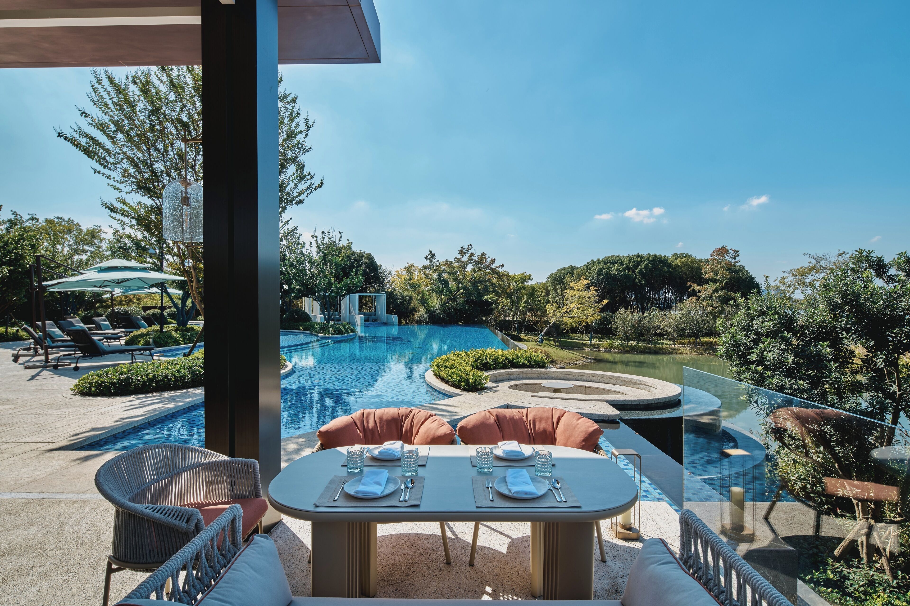 A poolside dining area with a table set for two, surrounded by wicker chairs and a glass-enclosed pool.