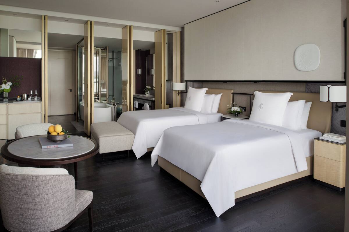 A modern hotel room with two beds, a round table with a fruit bowl, a sofa, and a large mirror.