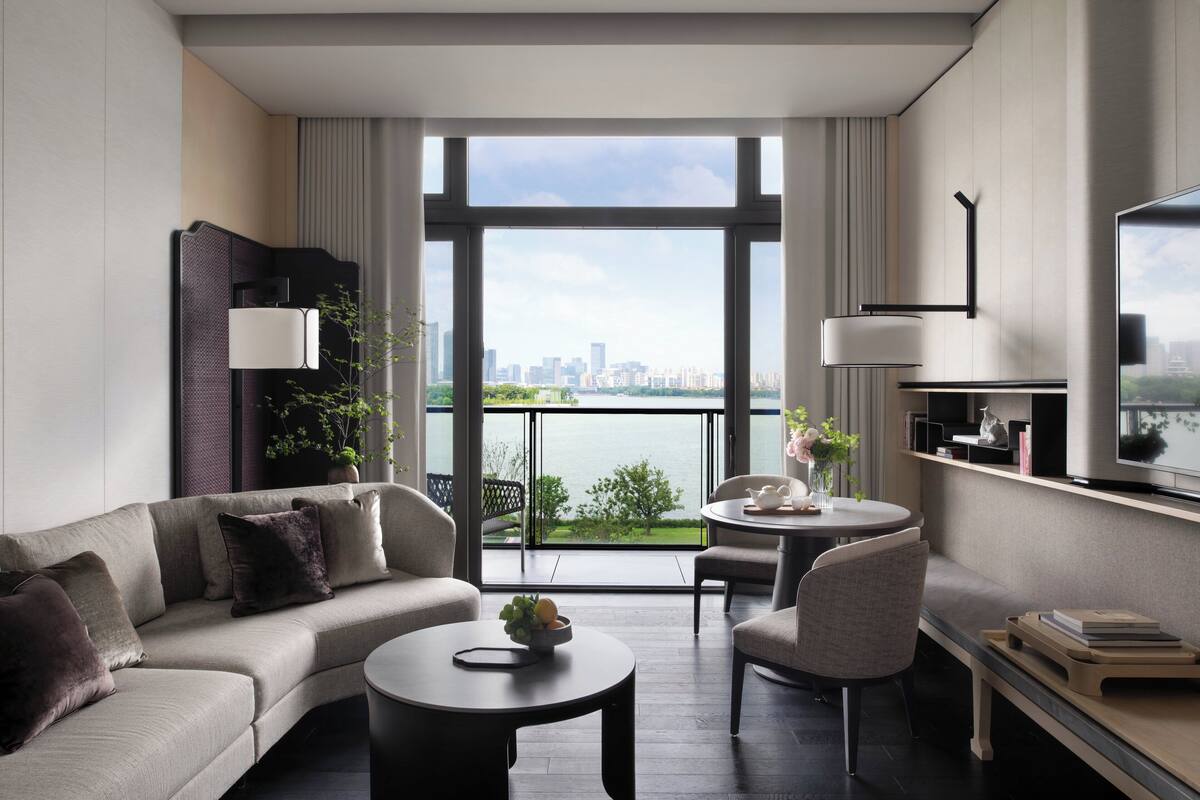 A modern hotel room with a sofa, a small round table, a chair, a TV, and a balcony with a city view.