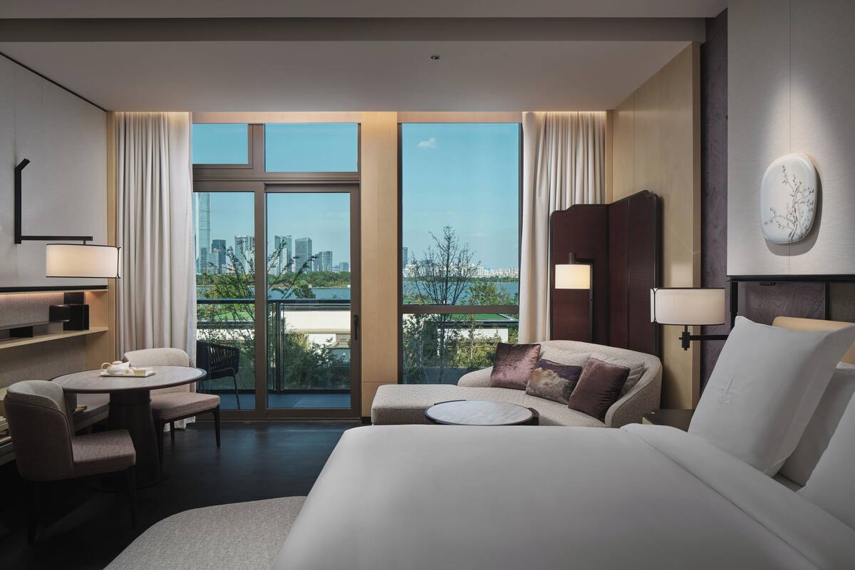 A modern hotel room with a large bed, a sitting area with a table and chairs, a view of the city through large windows, and a balcony.
