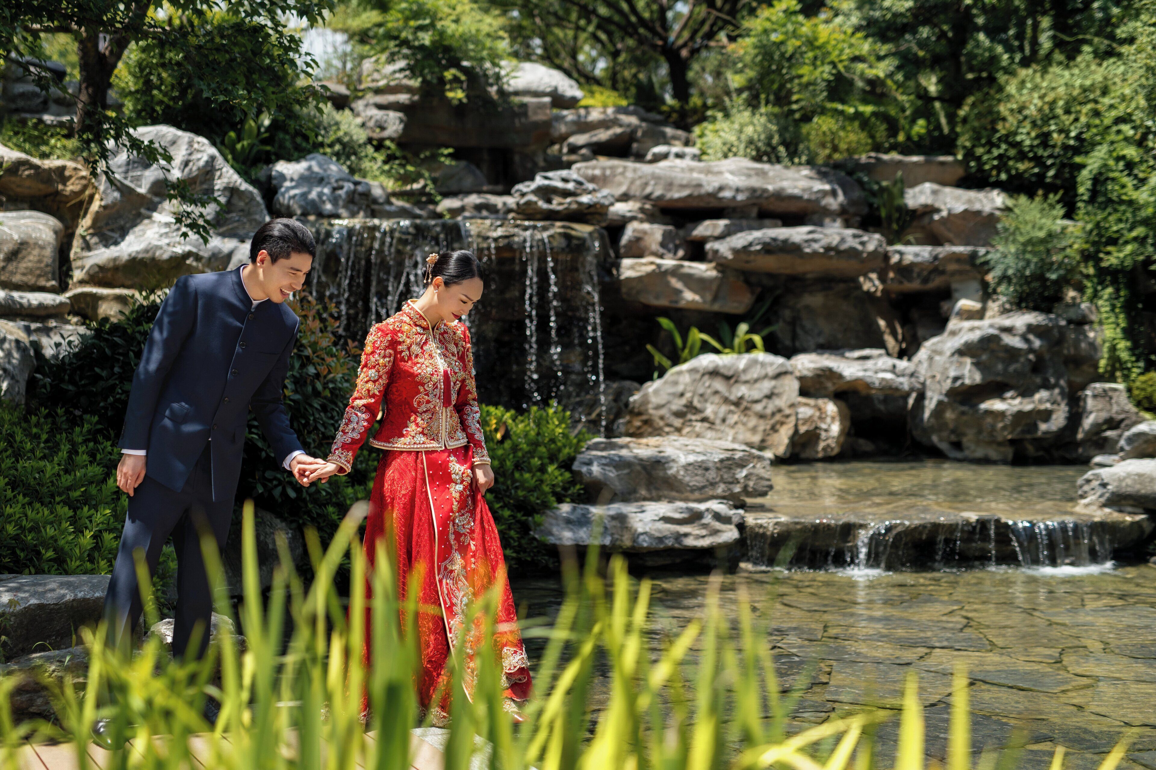 A couple walking near a waterfall in a garden with rocks and greenery.