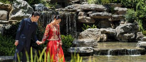 A couple walking near a waterfall in a garden with rocks and greenery.