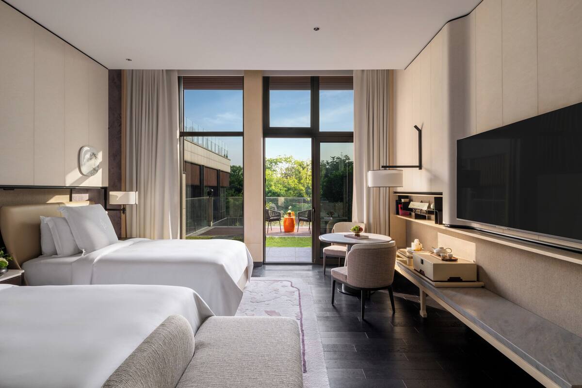 A hotel room with two beds, a TV, a desk with a chair, and a view of the outdoors.