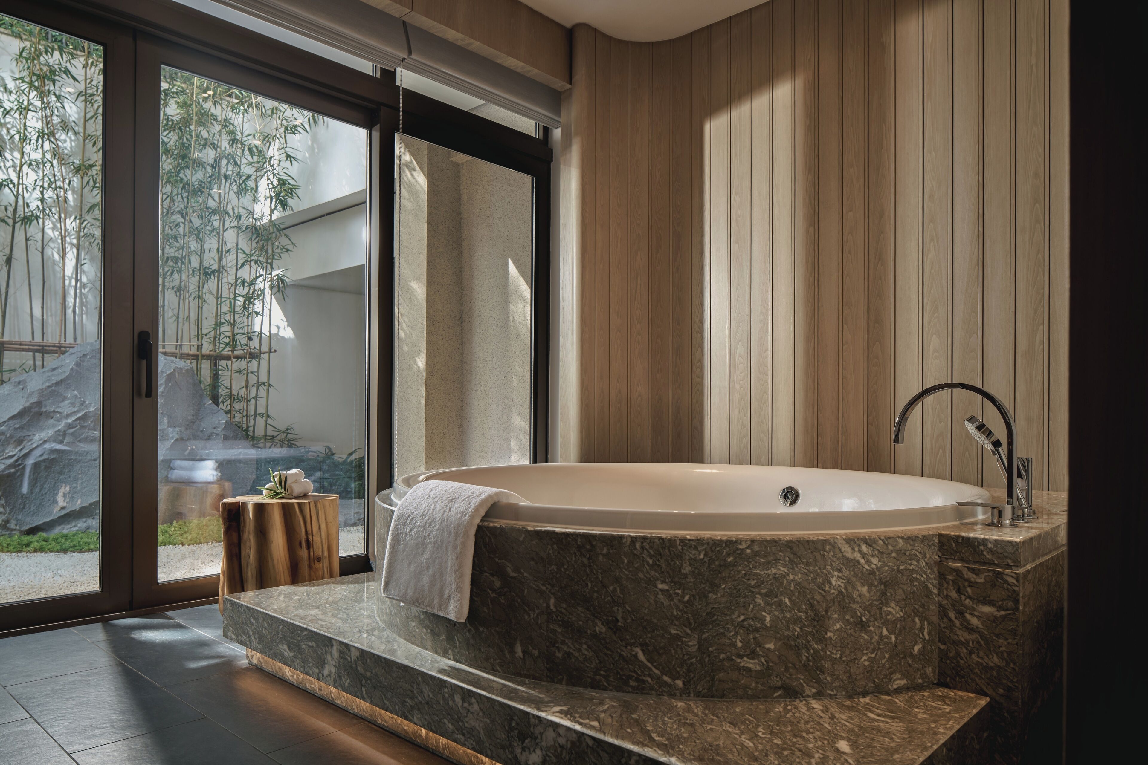 A modern bathroom with a freestanding bathtub, a wooden vanity, and a glass door leading to a garden.