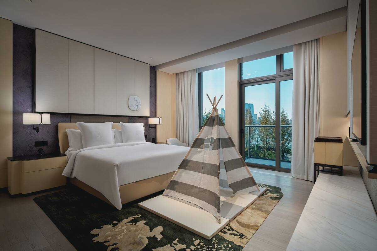 A modern hotel room with a large bed, a view of the city, and a teepee-like structure in the center.