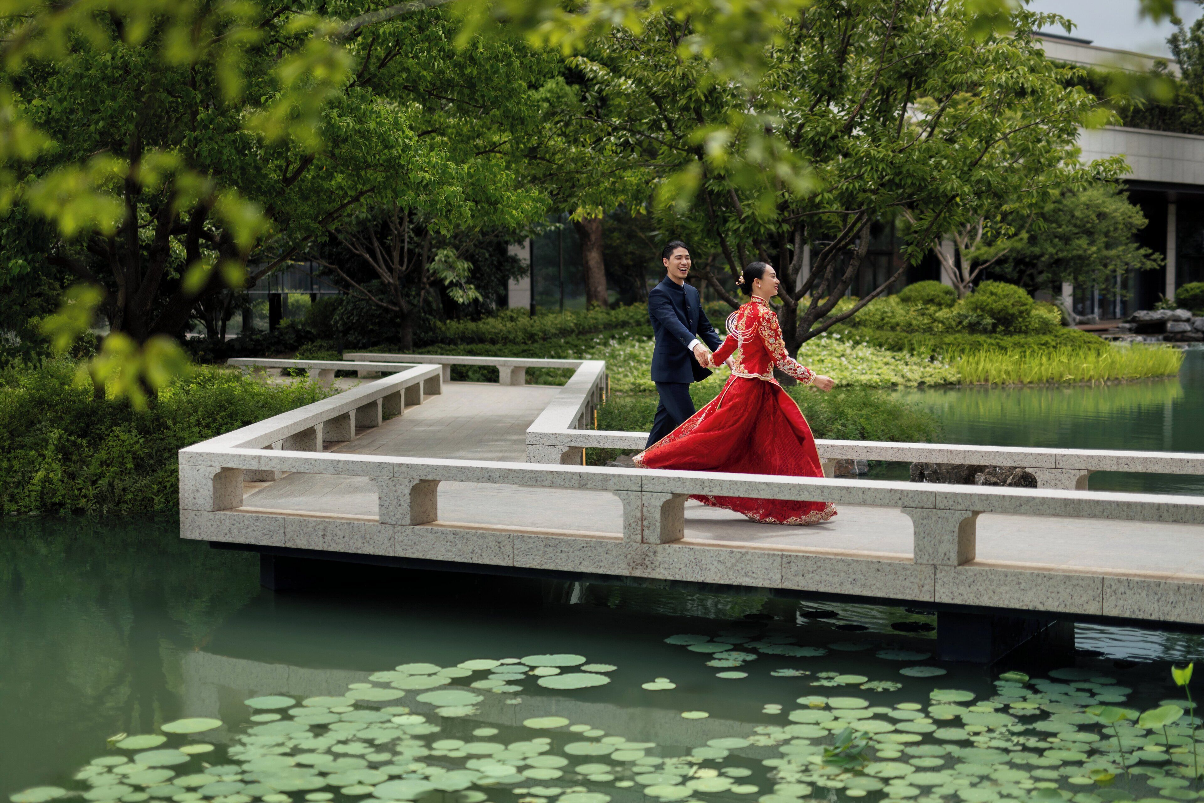 A couple walking on a stone bridge over a pond with lily pads.
