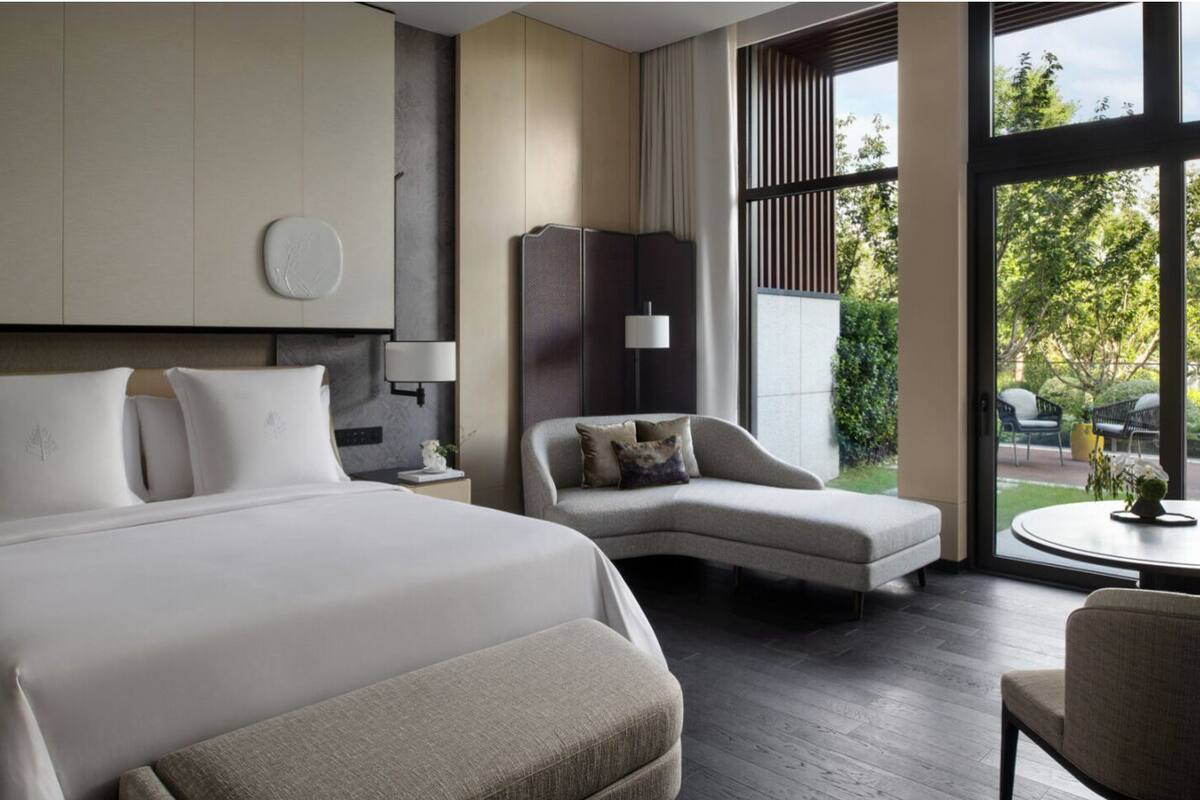 A modern hotel room with a large bed, a curved sofa, a small table, and a view of the outdoors.