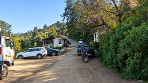 A dirt road with parked cars and a motorcycle, surrounded by greenery and a small building. - Buransh Heli Resort (Ukhimath)