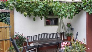 Un porche cubierto con una mesa y banco metálicos, rodeado de plantas en macetas y un portón de madera.
