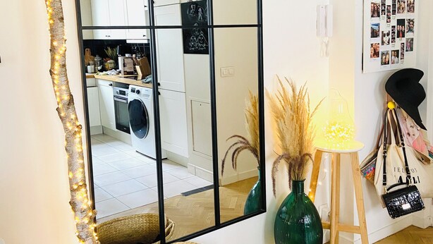 A room with a large mirror, a small table with a lamp, a vase with dried plants, and a wall with a photo collage. - Appartement Design Proche Paris & Versailles (Meudon)