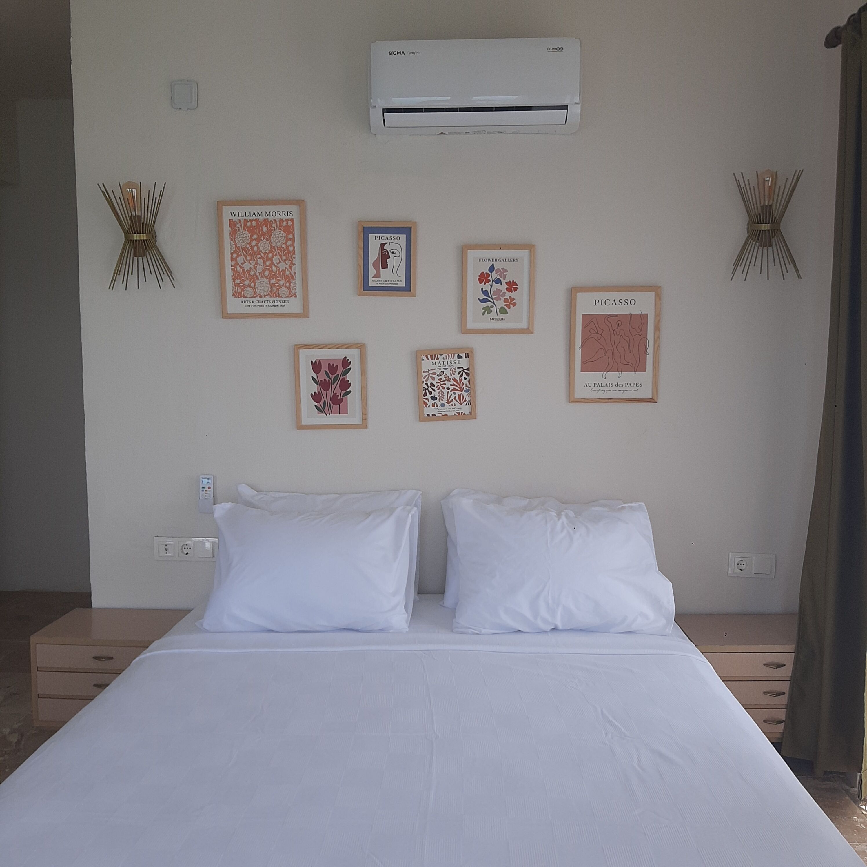 A hotel room with a bed, white pillows, a wall-mounted air conditioner, and several framed artworks including pieces by Picasso and Matisse.