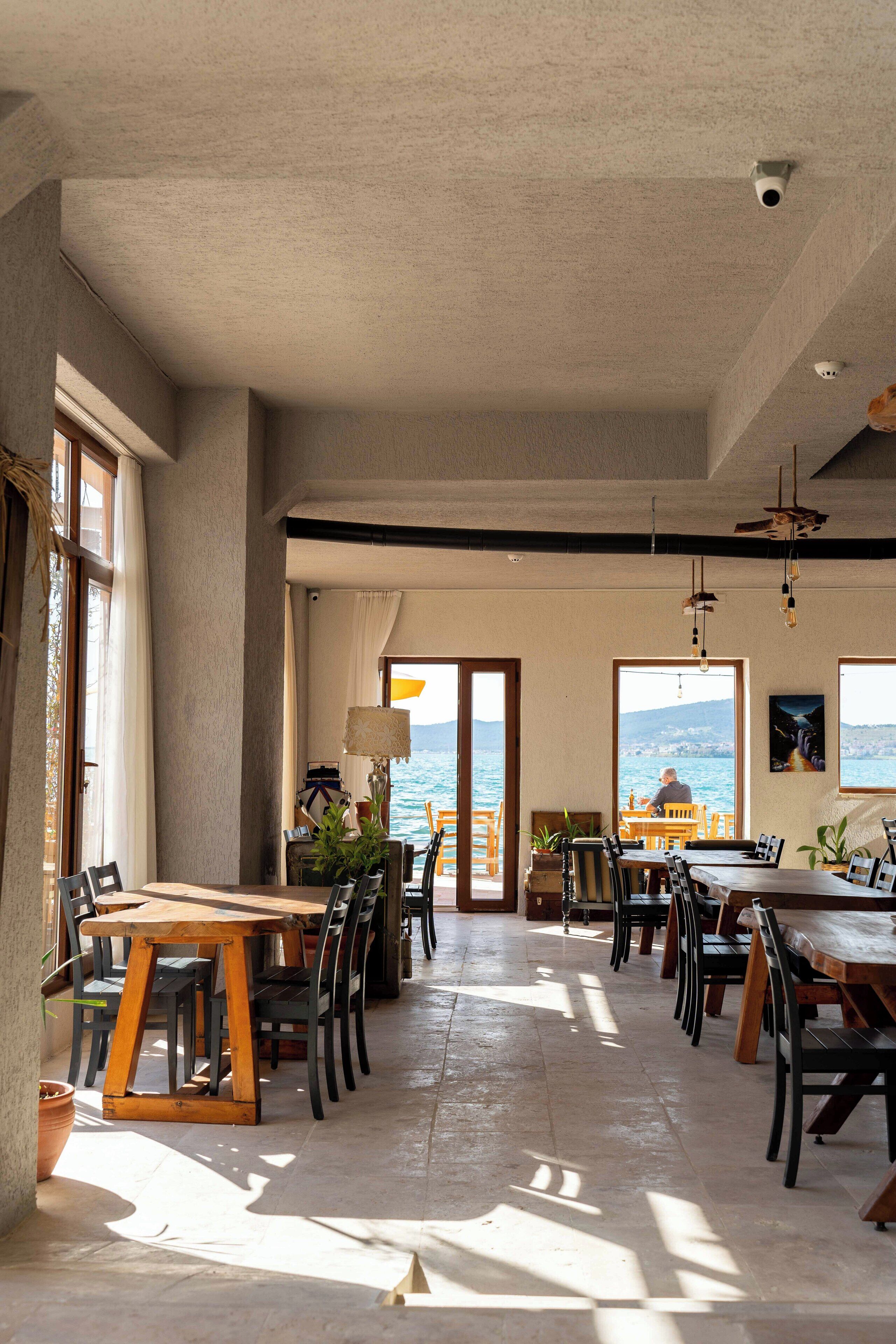 A spacious dining area with multiple tables and chairs, large windows offering a view of the sea, and a ceiling with a security camera.