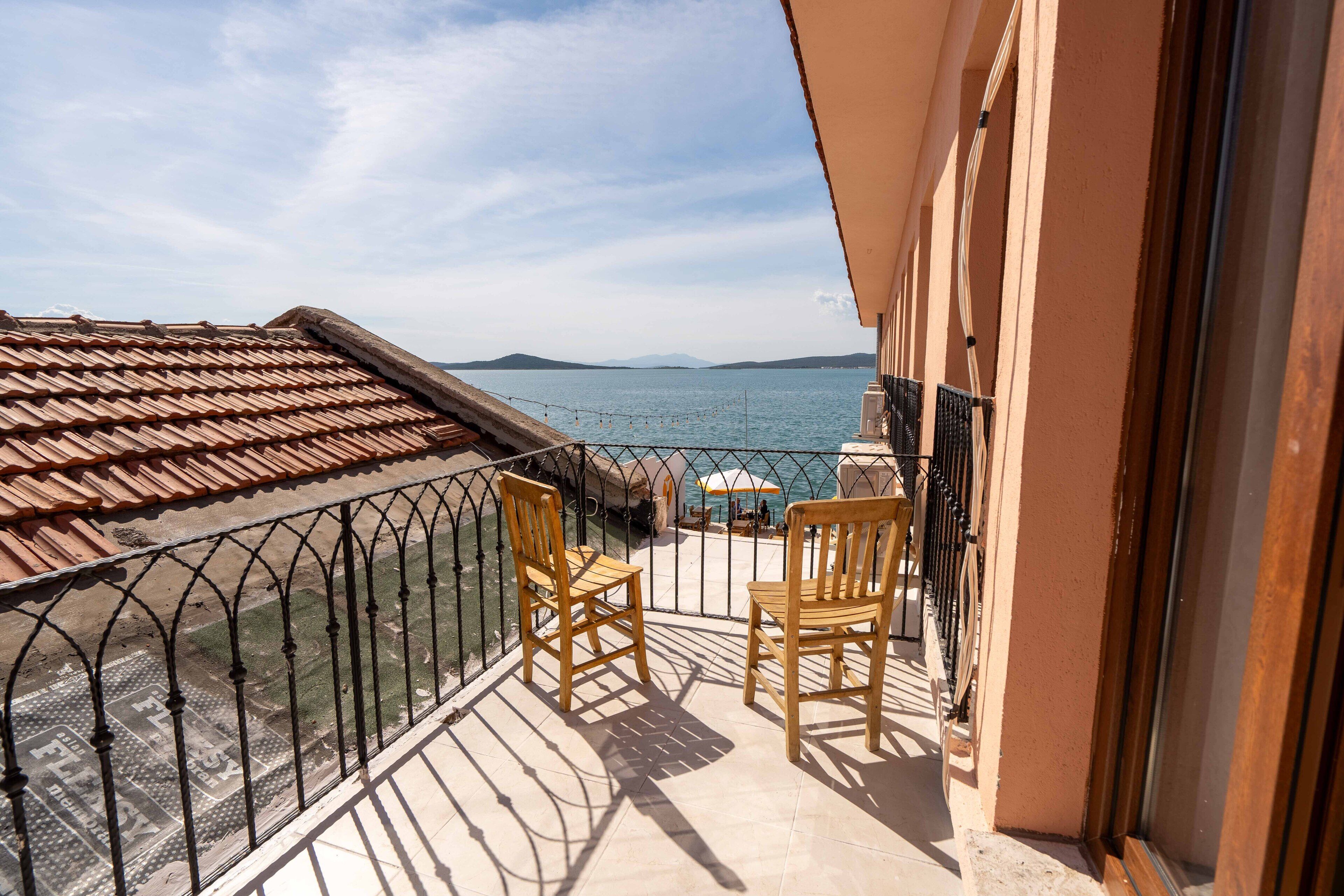 A balcony with a view of the sea, two wooden chairs, and a tiled floor.