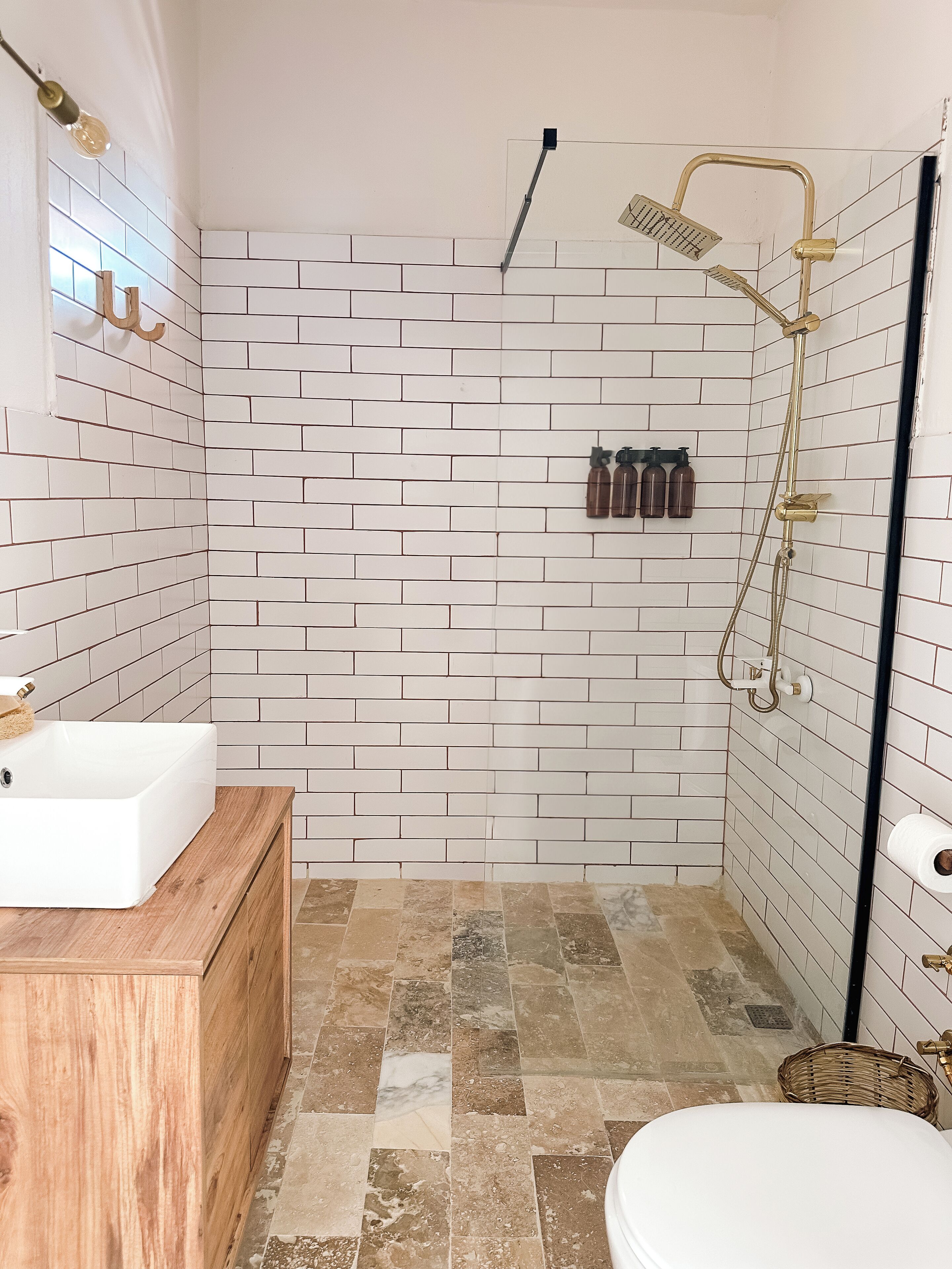 A bathroom with white brick walls, a wooden vanity, a white sink, a shower with a gold showerhead, and a toilet.