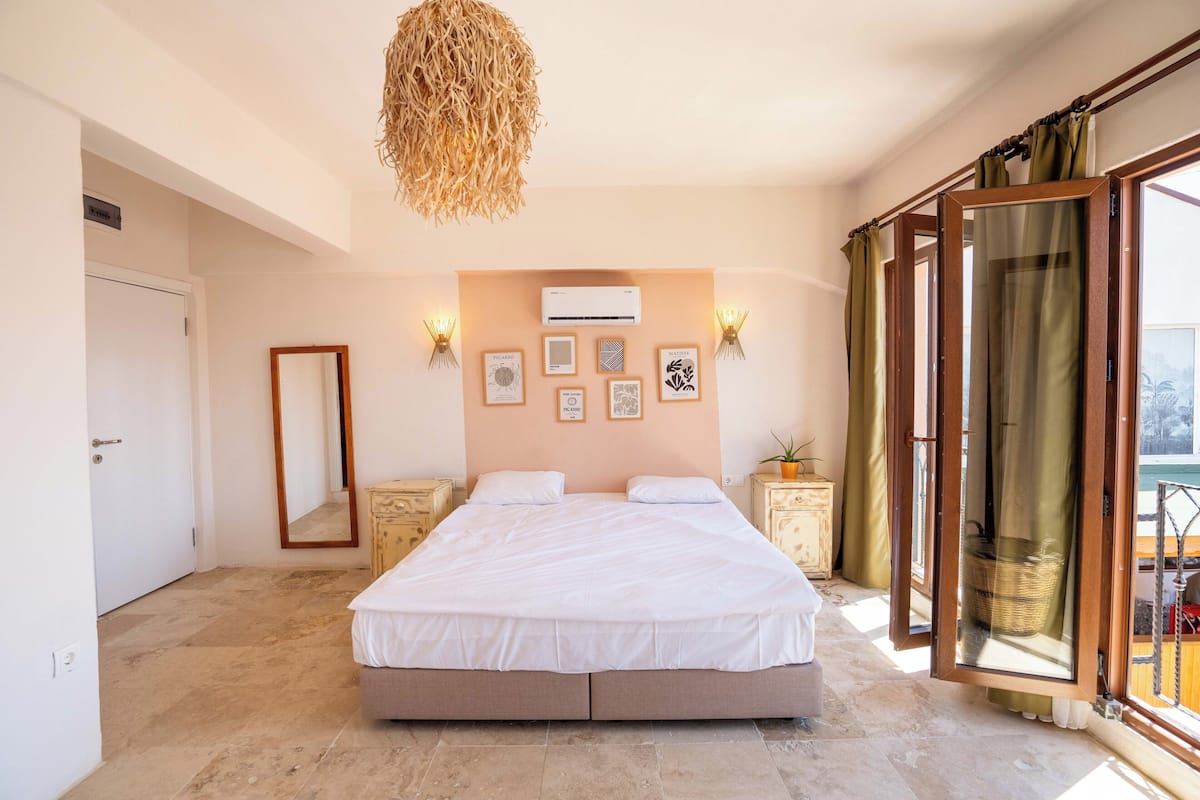 A bedroom with a large bed, a hanging light fixture, a wall-mounted air conditioner, and a window with curtains.