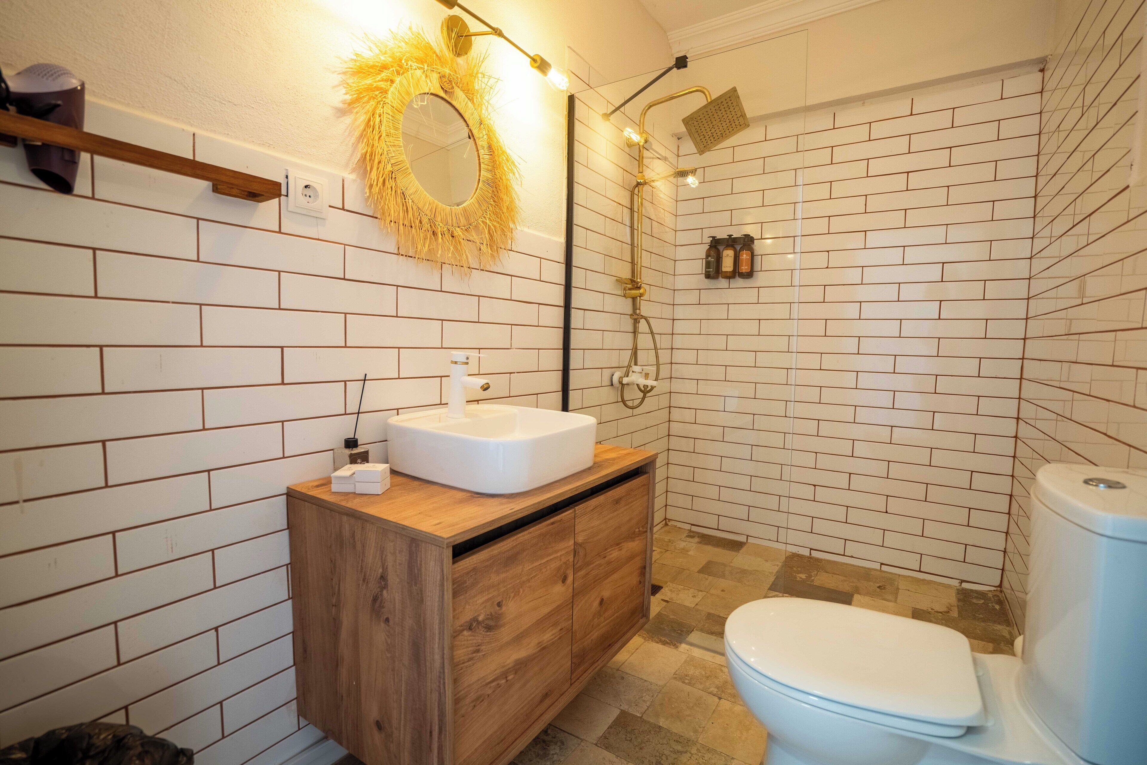Une salle de bain avec des murs en briques blanches, un meuble-lavabo en bois, un lavabo blanc, des toilettes et une douche dotée d’un pommeau de douche doré.