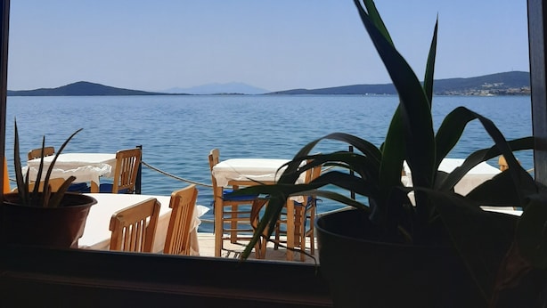 A view from a window overlooking a calm sea with a distant mountain range, wooden chairs, and potted plants on a terrace. - Casalyma Ayvalık (Ayvalik)