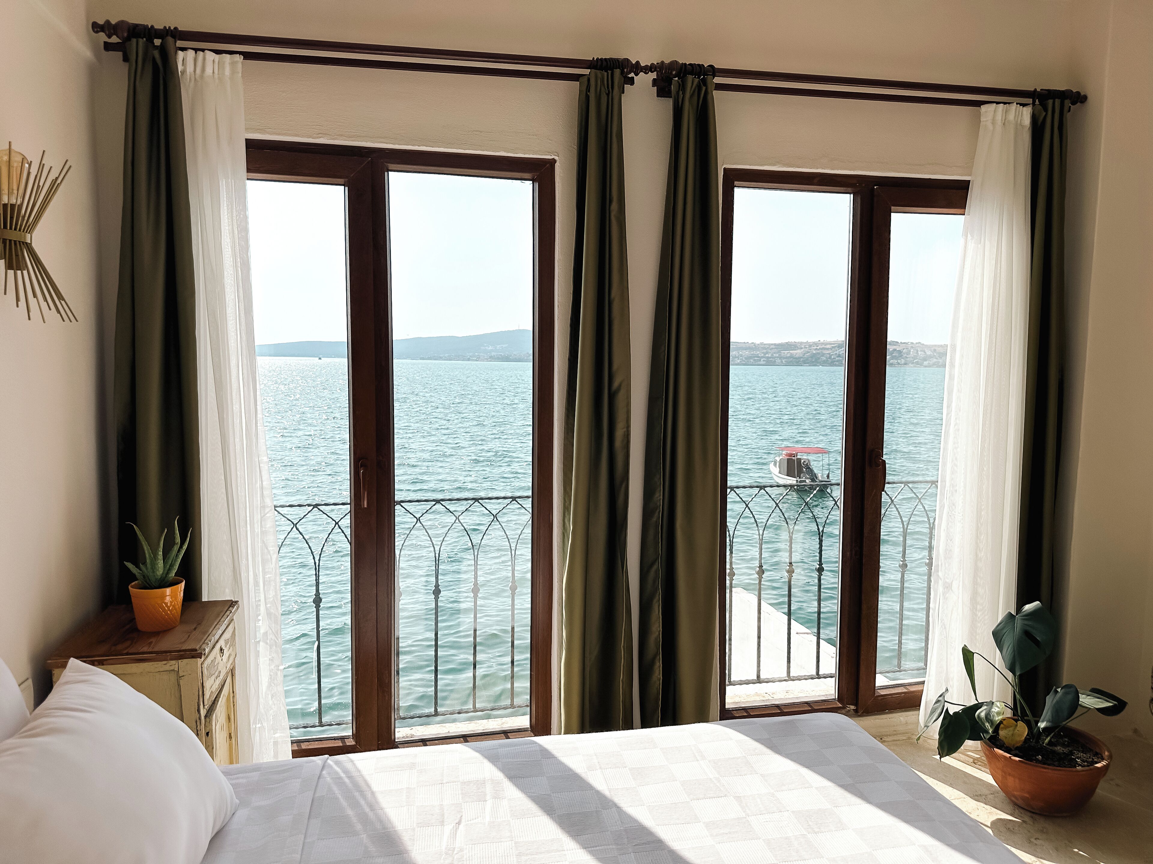 A bedroom with a bed, a potted plant, and a view of the sea through large windows.