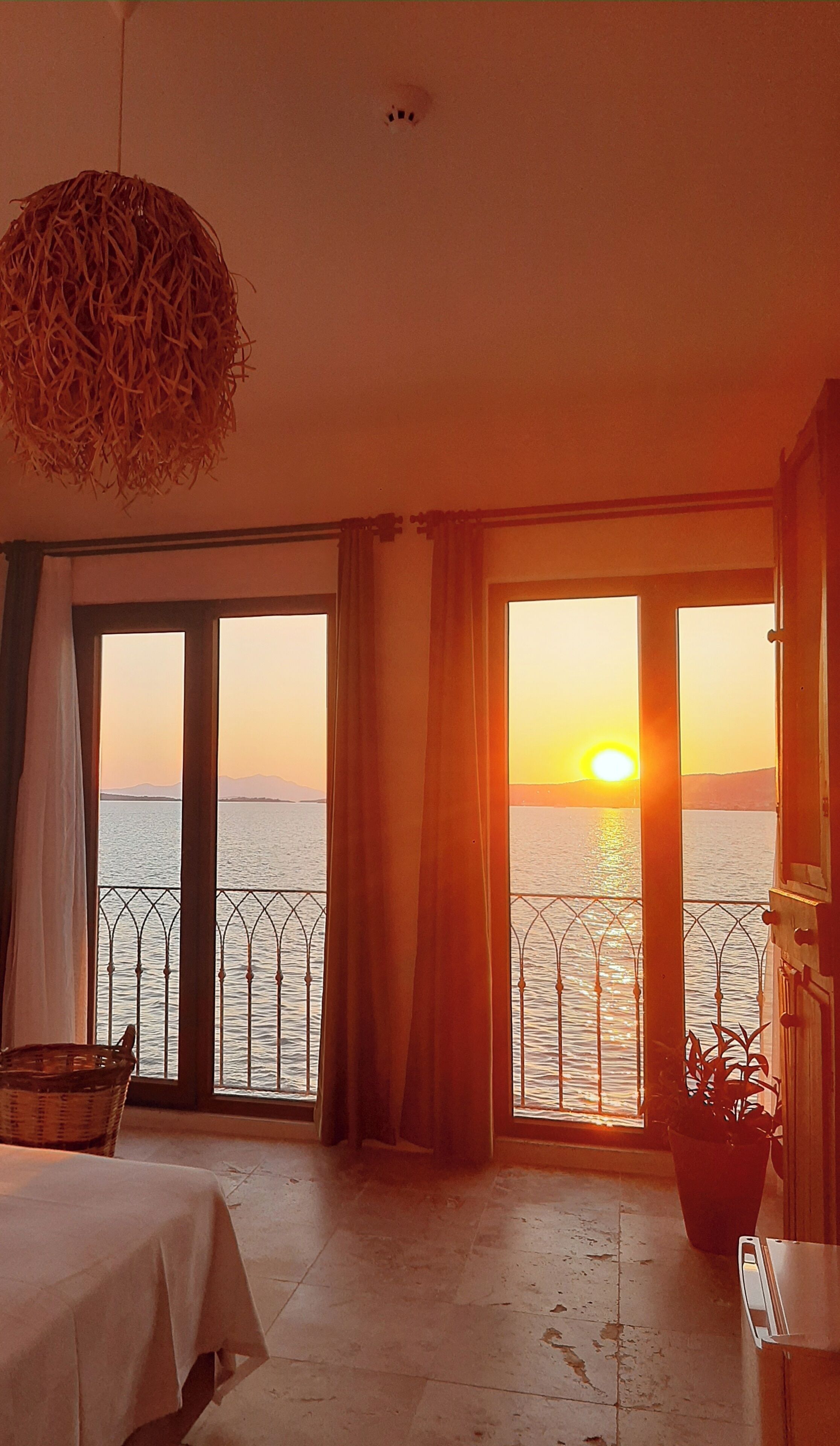 A room with a large window overlooking a body of water at sunset, a bed, a potted plant, and a hanging light fixture.