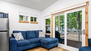 A modern living room with a blue sectional sofa, a built-in refrigerator, and a sliding glass door leading to an outdoor deck. - Hot Tub| Sauna| Arbutus Lake| Pines| 15 min to TC! (Traverse City)