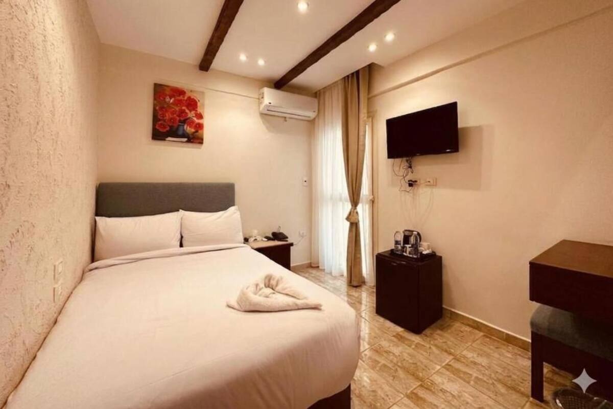 A hotel room with a bed, a flat-screen TV mounted on the wall, a bedside table with a lamp, and a window with curtains.