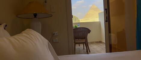 A hotel room with a bed, a lamp, a table with a chair, and a view of the pyramid.