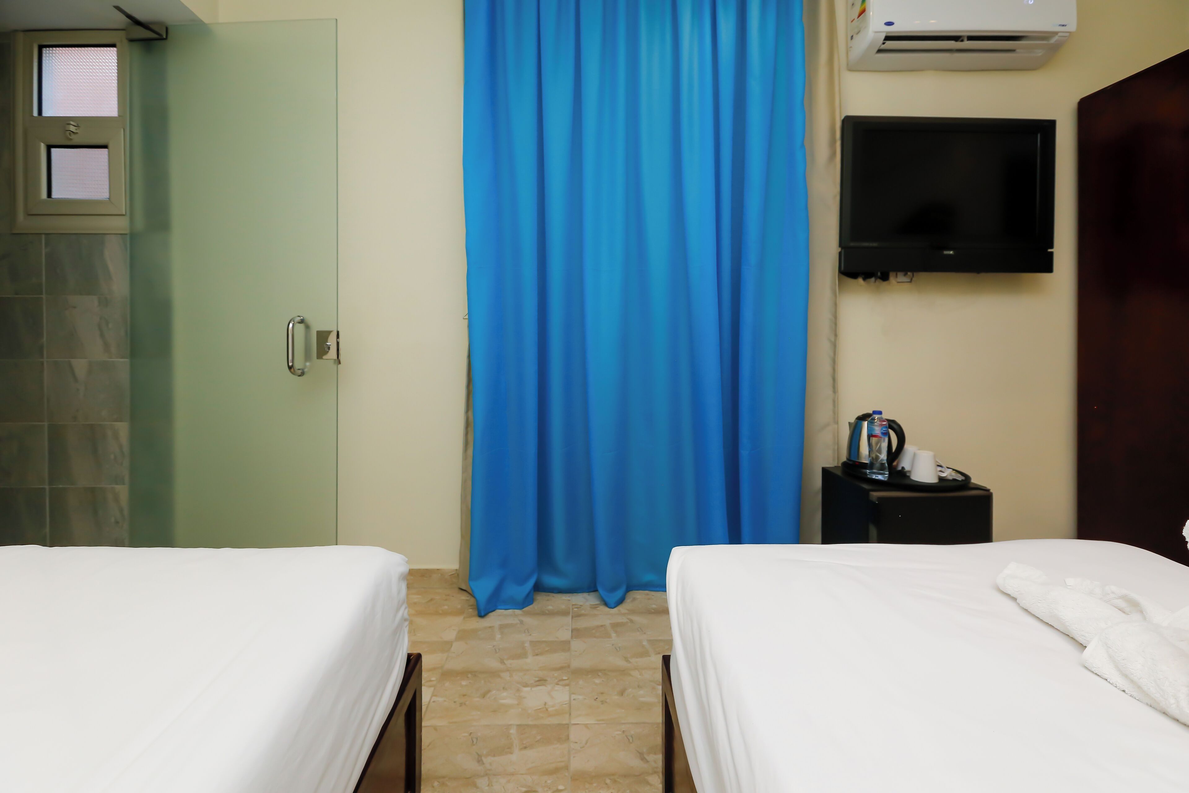 A hotel room with two beds, a blue curtain, an air conditioning unit, a TV, and a small table with a cup and a bottle.