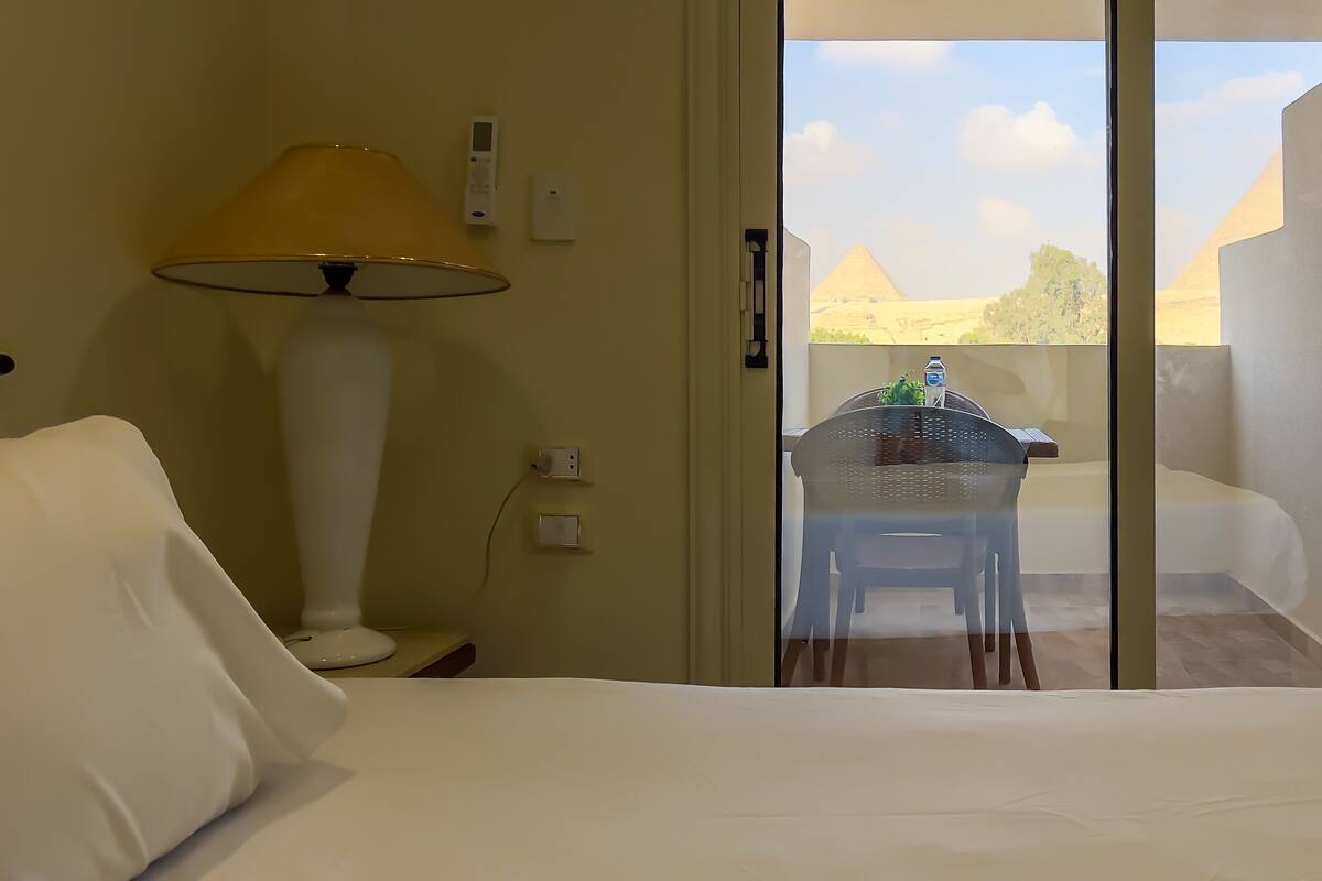 A hotel room with a bed, a desk, a lamp, and a view of the pyramids through a glass door.