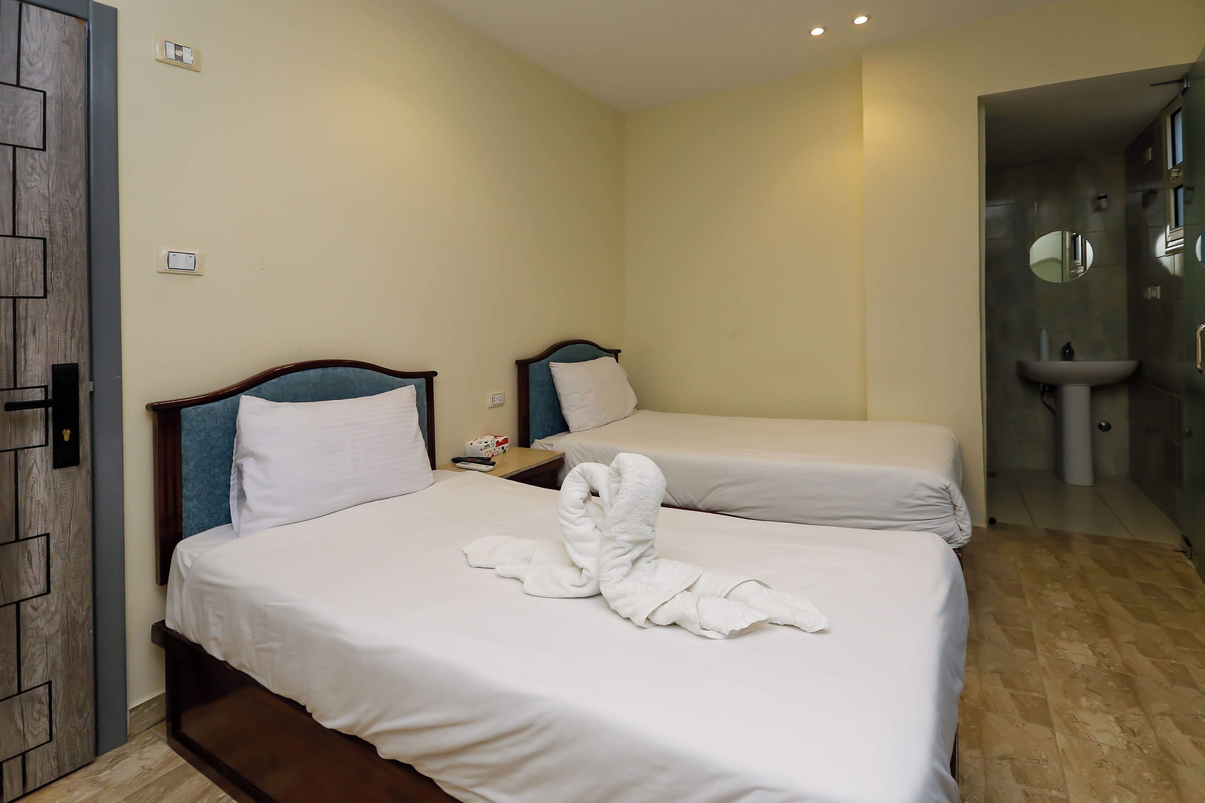 A hotel room with two beds, a wooden door, a small table with a lamp, and a bathroom with a sink and mirror.