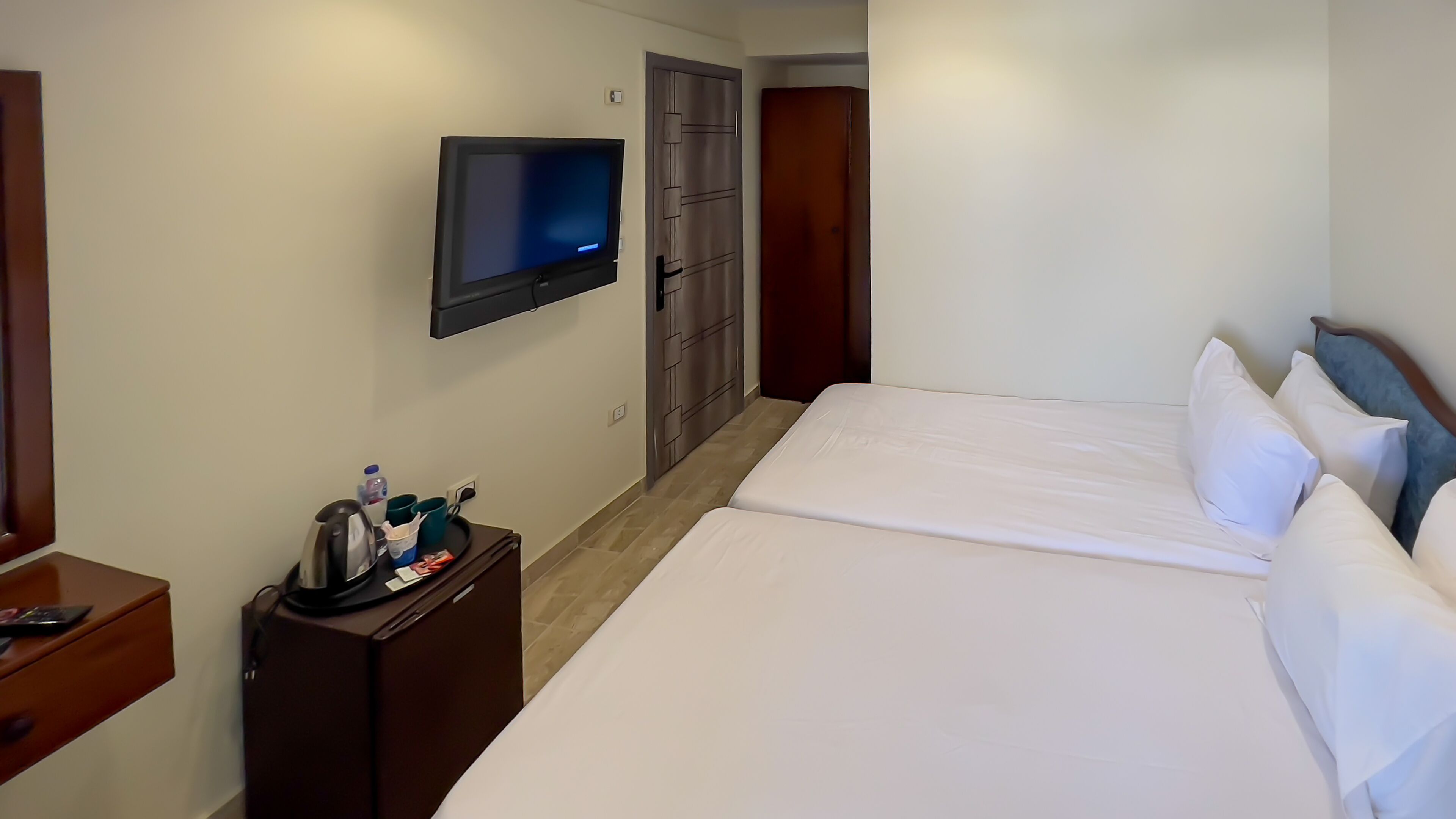 A hotel room with a bed, a TV mounted on the wall, a wooden nightstand, and a door.