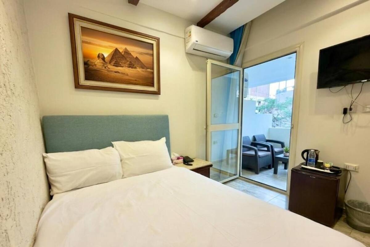 A hotel room with a bed, a painting of pyramids, a TV, and a sliding door to a balcony with a chair and a table.