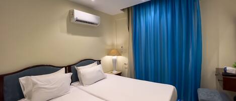 A hotel room with a bed, a bedside table with a lamp, an air conditioning unit, and a window with blue curtains.