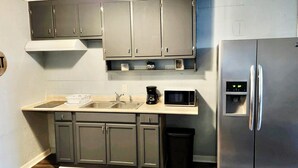 A kitchen with grey cabinets, a stainless steel refrigerator, a microwave, and a coffee maker on the countertop. - Greenhill Boutique Suites (Newport News)