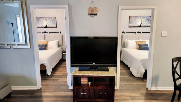A hotel room with a large flat-screen TV, two beds, a small wooden cabinet, and a mirror on the left wall. - Greenhill Boutique Suites (Newport News)