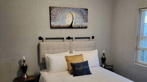 A bedroom with a bed, a nightstand, a lamp, a painting, and a window. - Greenhill Boutique Suites (Newport News)